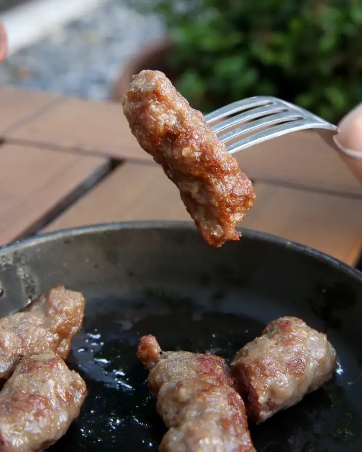 Komad junećeg ćevapa na viljušci iznad tiganja