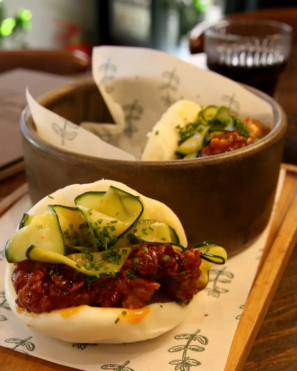 Pileći gua bao na drvenoj podlozi ispred kamene posude sa još jednim bao-om